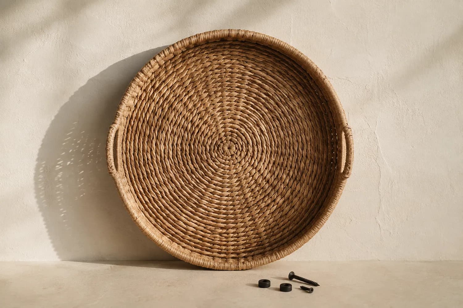 A flat woven wall basket held against a plain pale plaster wall, with two small wall anchors and screws lying on the surface below it.