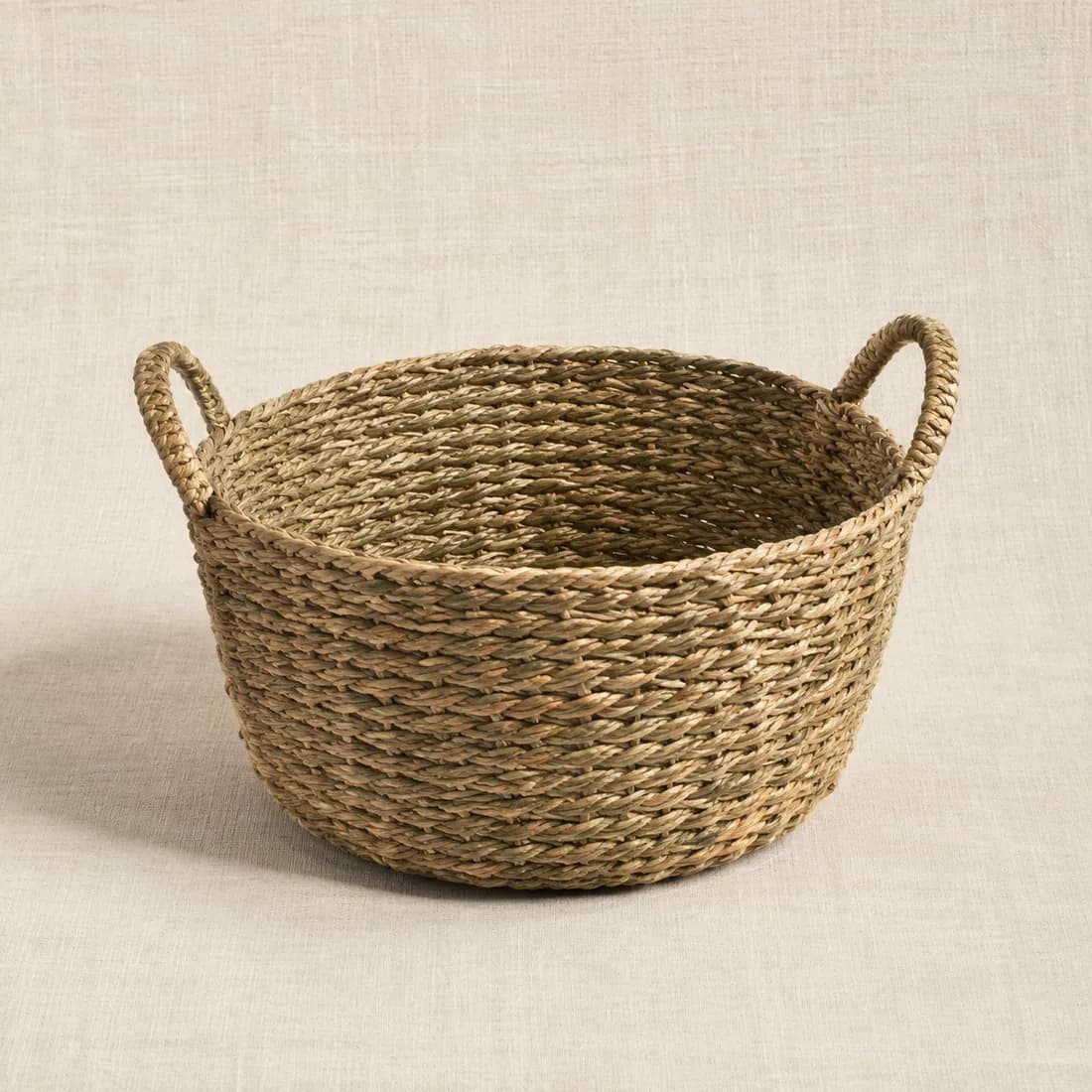 A round seagrass storage basket with two woven side grips, shown empty.